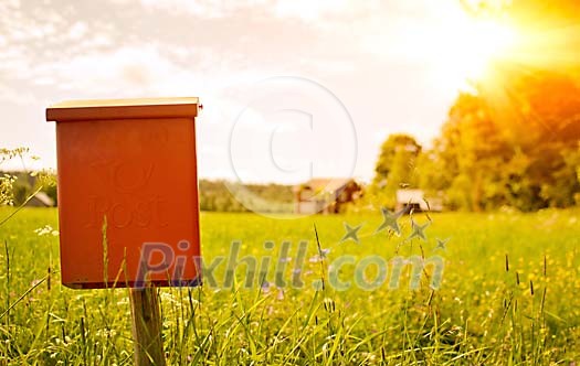 A postbox, mailbox with a house in the background and a sunset in summer. Green field