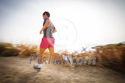 Caucasian young man running on a seacost path on a lovely summer evening, training for marathon (motion blurred image)