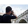 Portrait of a handsome young tourist taking photographs while sightseeing in Rome, Italy (with the Sant'Angelo bridge and castel in the background)