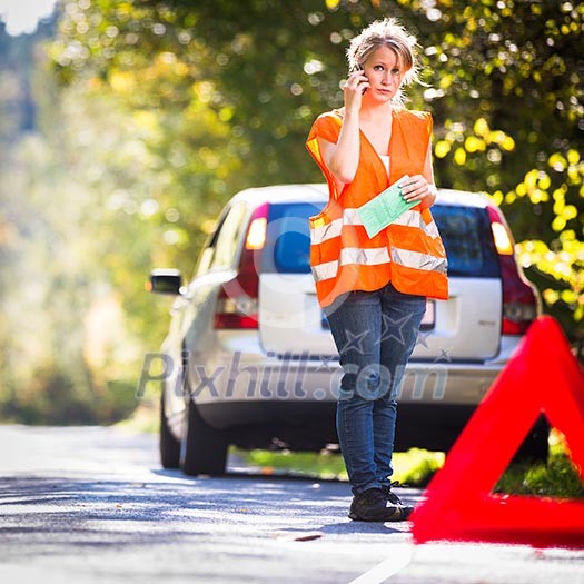 Young female driver wearing a high visibility vest, calling the roadside service/assistance after her car has broken down