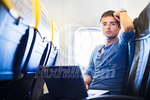 Handsome young man daydreaming while working on his laptop computer on board of an airplane during flight