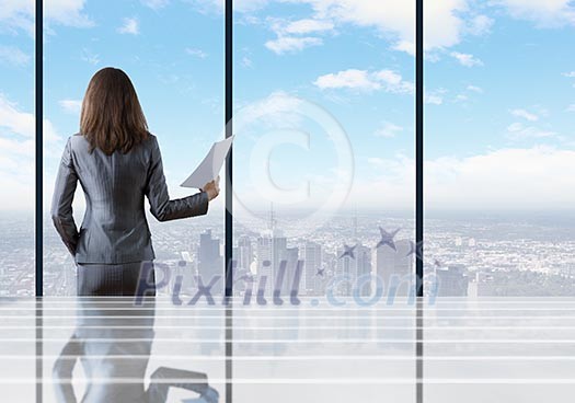 Businesswoman standing with back against office window holding documents in hand Businesswoman standing with back against office window holding documents in hand