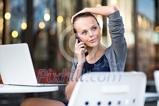 Portrait of a sleek young woman calling on a smartphone and using her laptop in a an
urban/city context (shallow DOF; color toned image)
