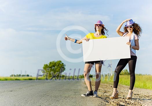Two pretty young girls standing aside road with blank white banner
