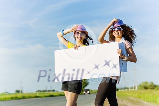 Two pretty young girls standing aside road with blank white banner Two pretty young girls standing aside road with blank white banner