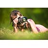 Pretty female photographer lying in grass, on a lovely summer day, taking pictures with her DSLR camera and a telephoto lens