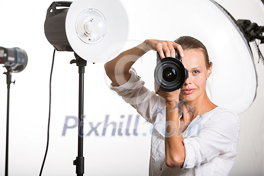 Pretty, female pro photographer with digital camera - DSLR and a huge telephoto lens in her well equiped studio, taking photos (color toned image; shallow DOF)