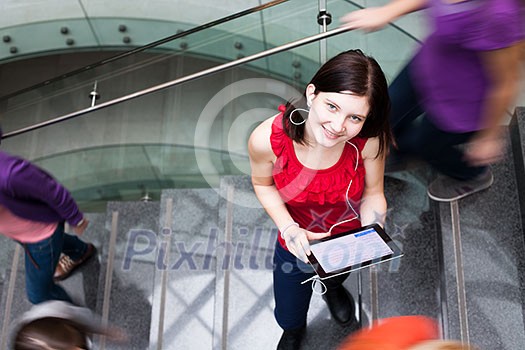 Pretty young student on the campus, holding a tablet computer while standing in the middle of a busy stairway, looking upwards