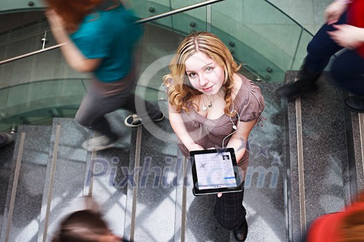 Pretty young student on the campus, holding a tablet computer while standing in the middle of a busy stairway, looking upwards