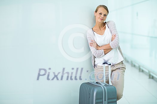 Pretty, young female passenger at the airport, waiting for her flight, wearing casual yet elegant clothes, arms folded, with a lovely smile and a big suitcase