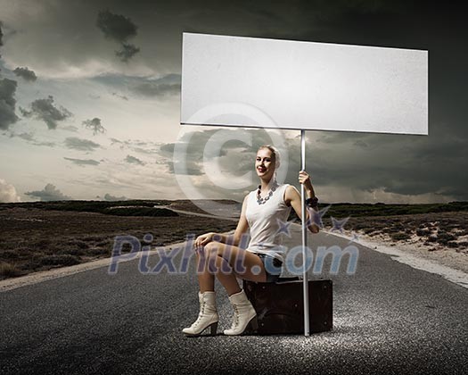 Young woman with blank banner sitting on suitcase