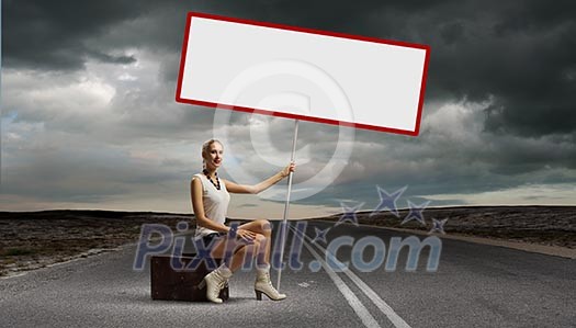 Young woman with blank banner sitting on suitcase