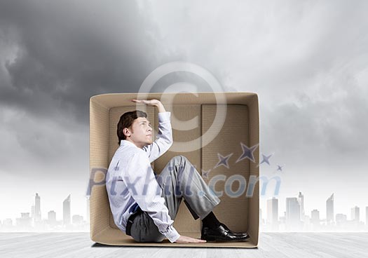 Young frustrated businessman sitting in small carton box