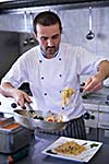 Handsome chef dressed in white uniform decorating pasta salad and seafood fish in modern kitchen