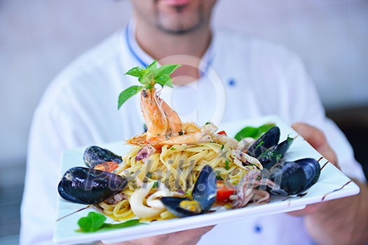 Handsome chef dressed in white uniform decorating pasta salad and seafood fish in modern kitchen