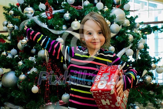 Smiling girl with giftbox looking at camera out of decorated firtree