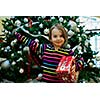 Smiling girl with giftbox looking at camera out of decorated firtree