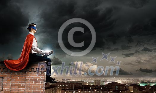Superman in cape and mask sitting on top of building and reading book Superman in cape and mask sitting on top of building and reading book