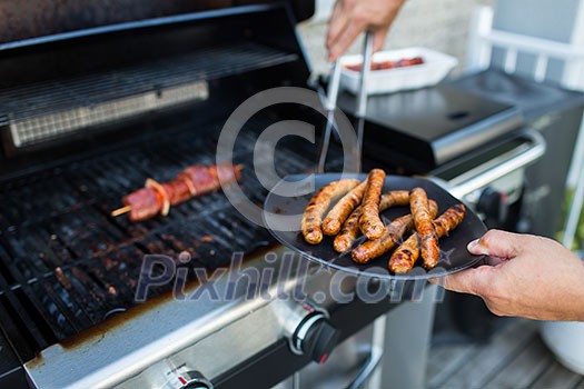 BBQ with sausages and red meat on the grill - male hands holding a plate and taking the meat off the grill before it is too late