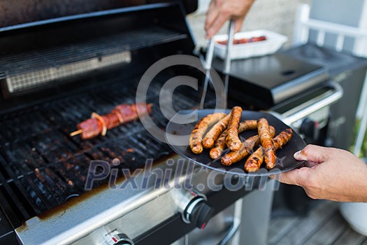 BBQ with sausages and red meat on the grill - male hands holding a plate and taking the meat off the grill before it is too late BBQ with sausages and red meat on the grill - male hands holding a plate and taking the meat off the grill before it is too late