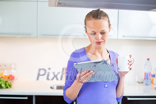 Pretty, young woman with her morning coffee and a tablet computer in modern kitchen - checking e-mails, reading news first thing in the morning