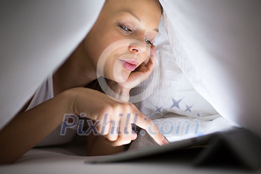 Pretty, young woman using her tablet computer in bed late at night, being online, browsing through the internet, reading news. staying in touch with friends, shopping online