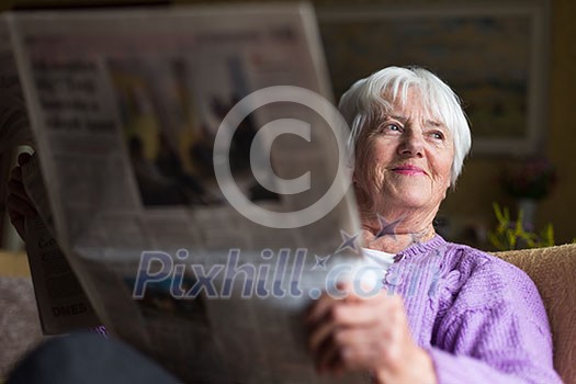 Senior woman reading morning newspaper, sitting in her favorite chair in her living room, looking happy