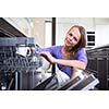 Pretty, young woman in her modern and well equiped kitchen putting cups into the dishwasher - an appliance that helps her keep the home tidy