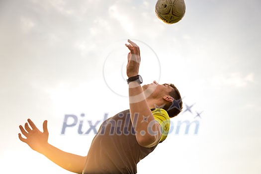 Young, male man playing soccer (lit by warm evening sunlight) heading the ball in the air