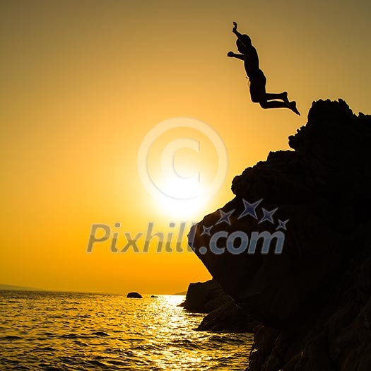 Boy jumping to the sea. Silhouette shot against the sunset sky. Boy jumping off a cliff into the ocean. Summer fun lifestyle.