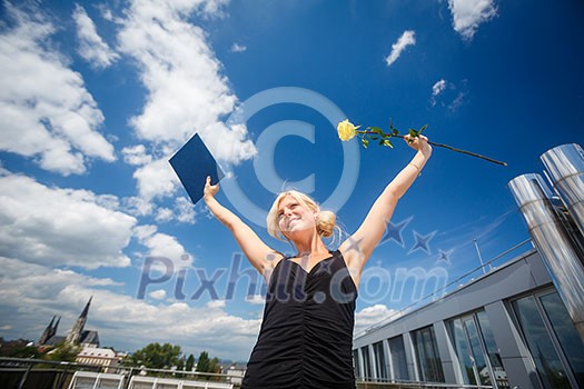 Pretty, young woman celebrating joyfully her graduation - spreading wide her arms, holding her diploma, savouring her success (color toned image; shallow DOF)