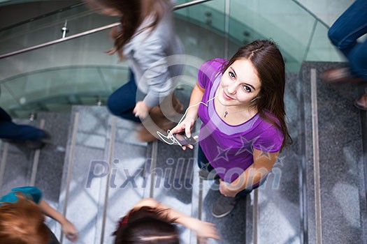 At the university/college - Students rushing up and down a busy stairway - confident pretty young female student looking upwards while listening to music on her mp3 player (color toned image) At the university/college - Students rushing up and down a busy stairway - confident pretty young female student looking upwards while listening to music on her mp3 player (color toned image)