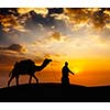 Rajasthan travel background - indian cameleer camel driver with camel silhouette in dunes of Thar desert on sunset. Jaisalmer, Rajasthan, India