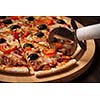 Pizza cutter wheel slicing ham pizza with capsicum and olives on wooden board on table