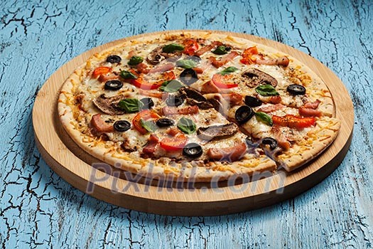 Ham pizza with capsicum, mushrooms, olives and basil leaves on wooden board on blue table Ham pizza with capsicum, mushrooms, olives and basil leaves on wooden board on blue table