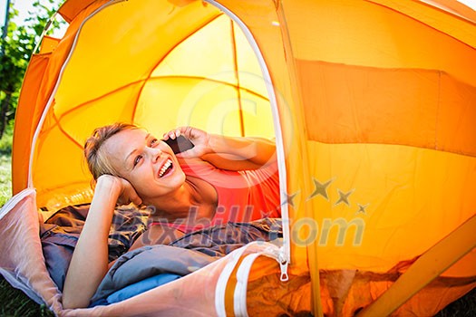 Pretty, young woman camping outdoors, lying in the tent in the morning, using her smart phone
