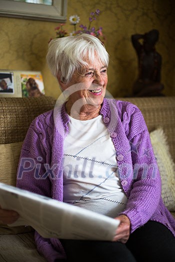 Senior woman reading morning newspaper, sitting in her favorite chair in her living room, looking happy