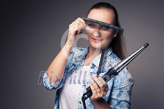 Funny portrait of a young female hairstylist, holding her tools and posing with a comb in front of her eyes ion front of a grey backdrop (color toned image; shallow DOF)