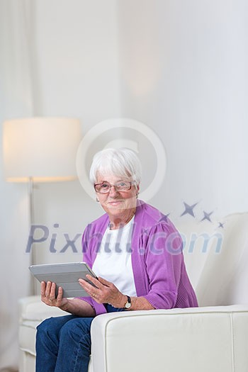 Portrait of a senior woman at home using a tablet computer - Looking happy, looking at the camera, smiling while sitting on the sofa in her living room