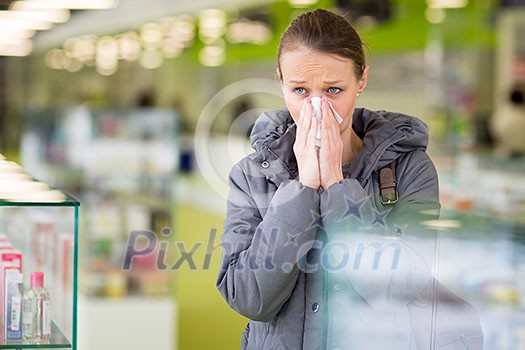 Young woman blowing her nose while in a modern pharmacy, choosing pills to help her with the flu/cold (shallow DOF; color toned image)