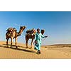 Rajasthan travel background - Indian cameleer (camel driver) with camels in dunes of Thar desert. Jaisalmer, Rajasthan, India