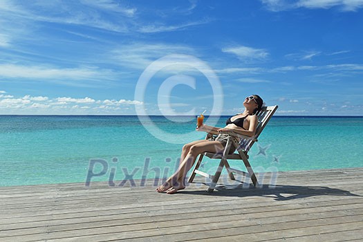 Beautiful young woman in bikini lying on a deckchair with a drink by the sea