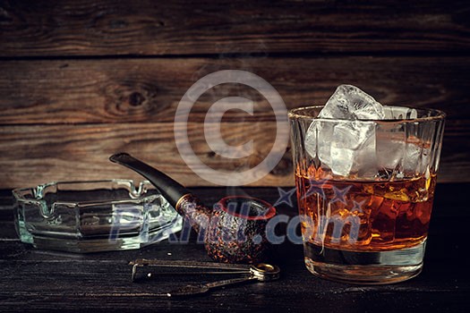 Glass of whiskey with ice and pipe on a wooden background. Vintage toned. Glass of whiskey with ice and pipe on a wooden background. Vintage toned.
