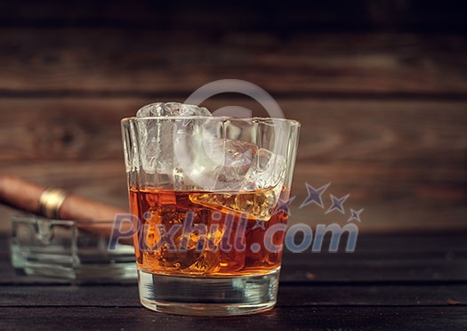 Glass of whiskey with ice and cigar on a wooden background. Vitage toned.