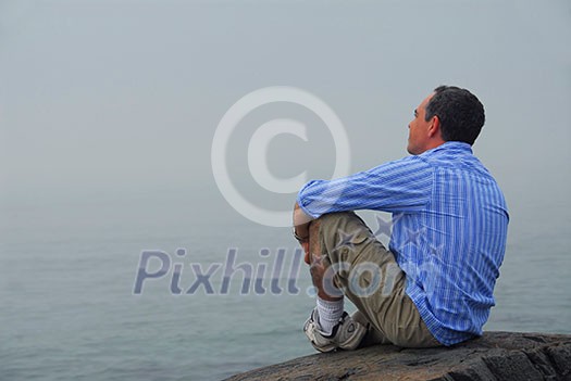 Man sitting on a rocky shore, looking at the foggy ocean. Uncertain future concept