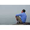 Man sitting on a rocky shore, looking at the foggy ocean. Uncertain future concept