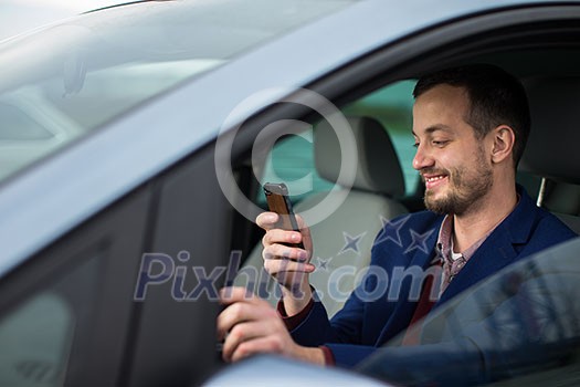 Handsome young man looking at his cellphone while at the wheel of his car, smiling