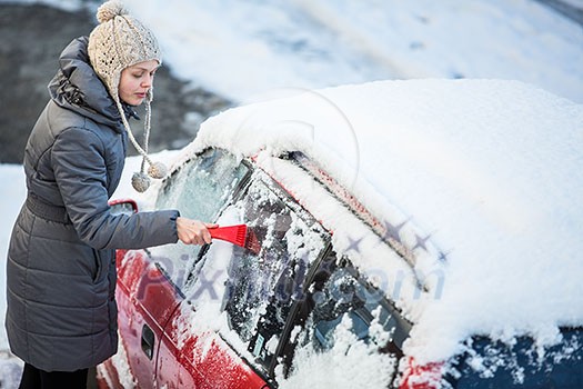 Young woman cleaning her car from snow and frost on a winter morning, she is freezing and needs to get to work