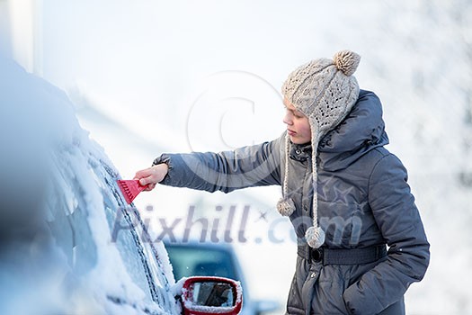 Young woman cleaning her car from snow and frost on a winter morning, she is freezing and needs to get to work