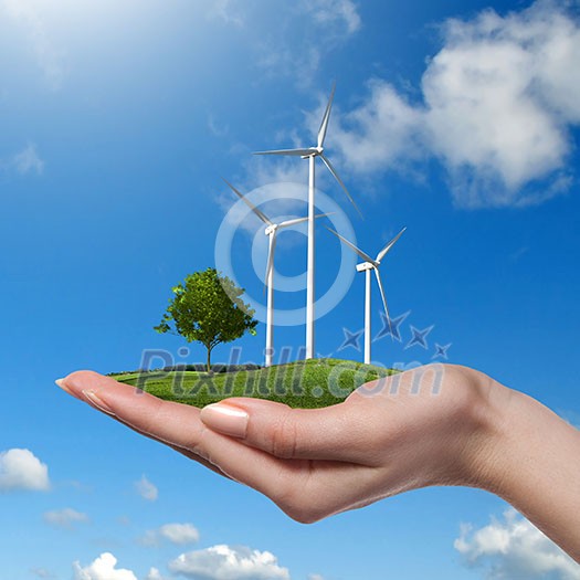 Wind turbines on meadow with tree holds in womans hand against blue sky and clouds. Green energy concept Wind turbines on meadow with tree holds in womans hand against blue sky and clouds. Green energy concept